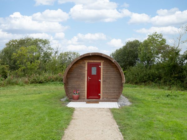 Small wooden cabin with a red door on a grassy field under a blue sky with clouds.