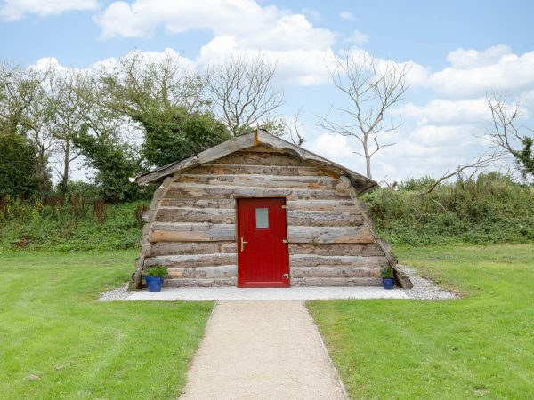 Small log cabin with a red door, set on green grass with trees and blue sky in the background.