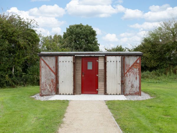 A shed with a red door and weathered metal walls stands on a grassy lawn under a blue sky.