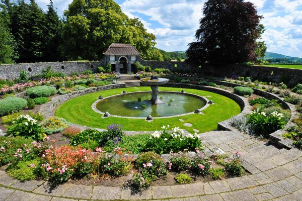 Circular garden with a central fountain, surrounded by flowers, greenery, and stone paths.