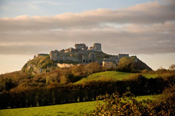 Ruins of a hilltop stone castle surrounded by green fields and under a cloudy sky.