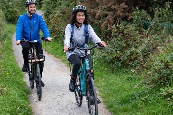 Two people wearing helmets ride bikes on a path surrounded by greenery and bushes.