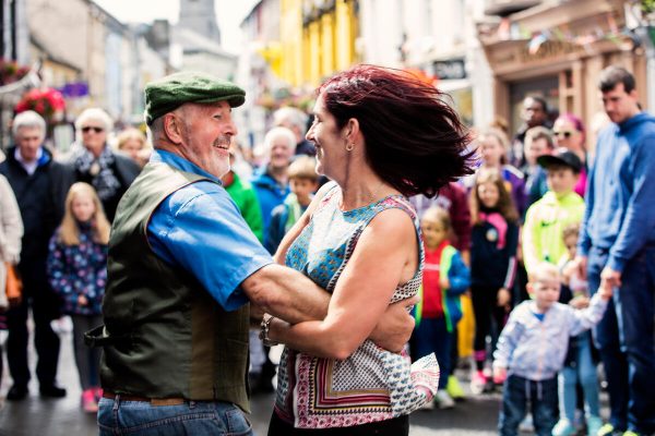 An older man and woman joyfully dance in a lively street surrounded by a crowd.