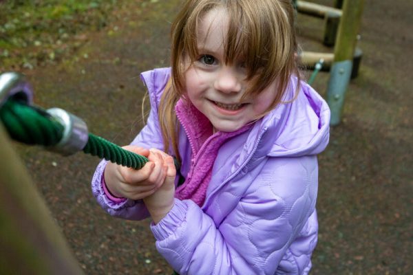 Smiling girl in a purple jacket holds onto a green rope at a playground.