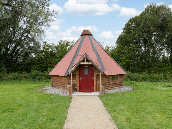 Small wooden cabin with a red roof and door, surrounded by grass and trees.