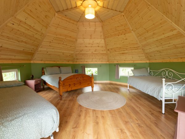 Spacious wooden room with three beds, round rug, and small windows around the walls.
