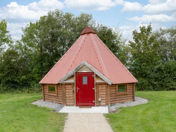 A small, round log cabin with a red door and cone-shaped red roof, set in a grassy area.